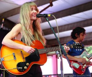 Female guitarist smiling while singing into a microphone on stage, playing an orange sunburst guitar.