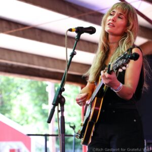Female musician singing into a microphone while playing an acoustic guitar on a stage outdoors.