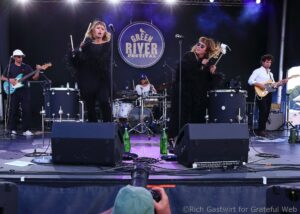 Band on stage at Green River Festival; two female singers in black feathered outfits with a drummer and guitarists on either side.
