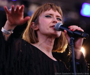 Close-up of a female singer performing into a microphone, with a raised hand and stage lights in the background.