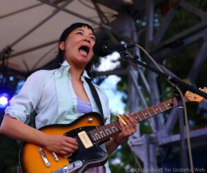 Female guitarist sings into a microphone on stage, wearing a light blue shirt and playing a sunburst electric guitar in a live outdoor performance.