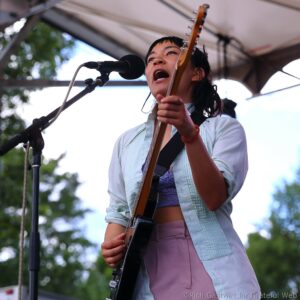 Female guitarist singing into a microphone during an outdoor live performance.