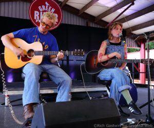Two musicians on a stage: a man in a blue shirt playing an acoustic guitar and a woman singing into a microphone with tattoos on her arms.