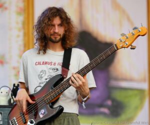 Musician with long curly hair playing a bass guitar on stage, wearing a light T-shirt and striped shorts