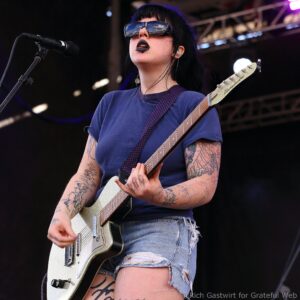 Female guitarist on stage wearing sunglasses and a blue shirt, playing a white electric guitar with a purple strap, showing lots of tattoos on arms and legs