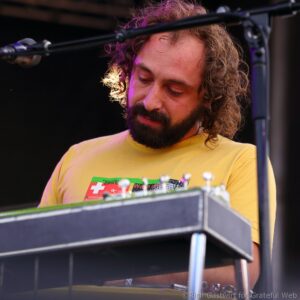 Bearded musician with long wavy hair in a yellow shirt on stage, viewed over a keyboard or instrument in the foreground.