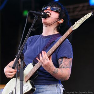 Musician singing into microphone while playing an electric guitar on stage, wearing sunglasses and a blue shirt, with tattooed arms visible.