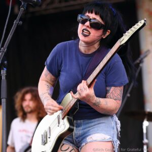 Female guitarist with black hair and sunglasses, wearing a blue shirt, playing a white electric guitar on stage