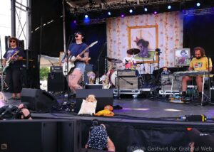 Band performs on a decorated outdoor stage with musicians, drums, and keyboards amid framed artwork backdrop.