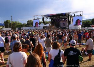 Large outdoor concert with a packed crowd facing a stage with two large video screens and a banner.