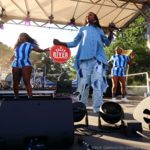 Three female performers sing and dance on an outdoor stage; a drummer is in the background with the 'Green River Festival' sign visible.
