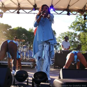 Female singer on an outdoor stage, pointing toward the camera while performing in a frayed blue denim dress, with band members in the background.