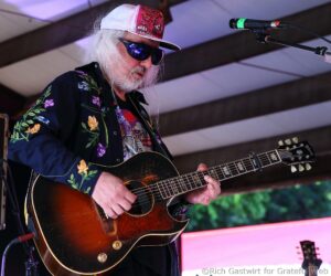 Guitarist with long white hair and a floral jacket playing a sunburst electric guitar on stage, wearing a white cap and sunglasses.