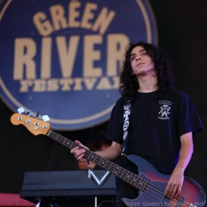 Musician playing a bass guitar on stage, with a large blue festival sign visible behind them.