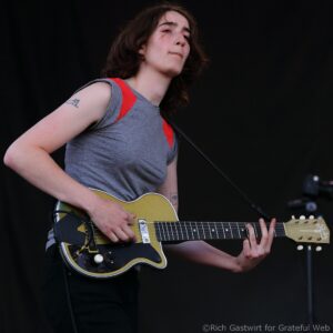 Musician playing an electric guitar on stage, left hand fretting the neck and right hand near the pickups, wearing a gray sleeveless shirt with red trim against a dark background.