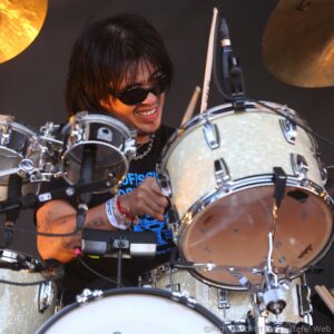 Drummer with dark hair and sunglasses performing on stage behind a silver drum kit with microphones nearby.