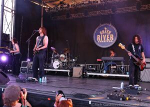 Band performs on an outdoor stage at Green River Festival; lead singer with guitar in front of microphone, drummer behind, guitarist on left.