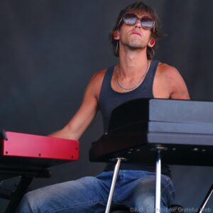 Musician wearing sunglasses and a dark tank top, seated at a red keyboard on stage, concentrating on playing.