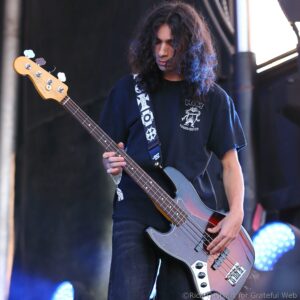 Male bassist with long hair playing a sunburst bass guitar on stage, stage lights in the background.