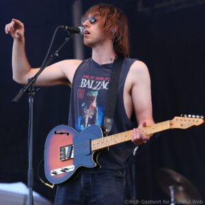 Guitarist singing into a microphone on stage while playing a sunburst electric guitar with a strap over a sleeveless shirt.