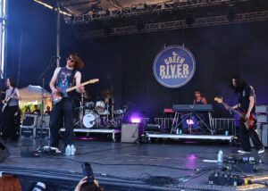Live rock band performing on an outdoor stage at the Green River Festival, guitarist at front with sunglasses and a Telecaster-style guitar, drummer behind them, big circular festival sign in the backdrop.
