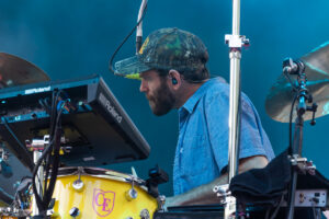 Drummer in a camo cap and blue denim shirt plays a Roland keyboard mounted above a yellow drum kit at a live performance.