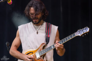 Male guitarist on stage playing a sunburst electric guitar; sleeveless beige shirt, colorful strap, long hair, and beard.
