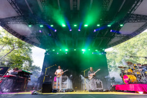 Outdoor concert stage with green lighting; two guitarists center and right, keyboardist left, drummer on a pink riser to the right, with lush trees in the background.