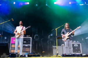 Two musicians perform on an outdoor stage under blue, green, and purple lights; a guitarist on the left sings while a bassist on the right plays.