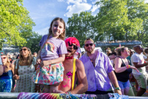Crowd at a pride festival with a shirtless man wearing a pink wig holding a young girl on his shoulders, both smiling, surrounded by cheering attendees.