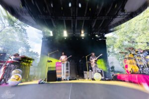 Two guitarists and a keyboardist perform on an outdoor festival stage with drums on the right and colorful lighting.