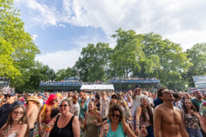 Crowd of people dancing and enjoying an outdoor summer concert in a park with banners and trees in the background.
