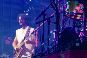 Guitarist with a beard playing an electric guitar on a stage under blue-purple lights, microphones and gear around him.