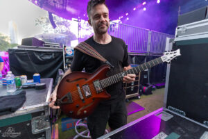 Guitarist in a black t-shirt playing a brown semi-hollow electric guitar backstage at a concert, purple stage lights in the background.