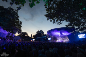 Outdoor concert at dusk with a large crowd watching a lit stage under trees in a park setting.