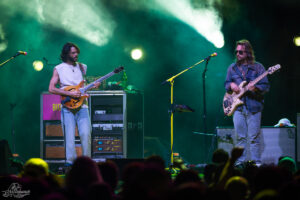 Two musicians performing live on stage with guitars, colorful lights, and a cheering crowd in the foreground.