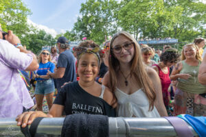 Two smiling young women pose behind a metal barrier at an outdoor festival, with a crowd and green trees in the background.