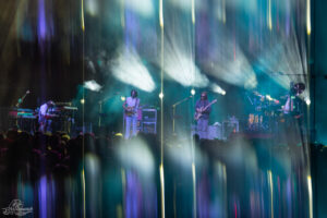 Live concert on stage with musicians playing guitar, bass, keyboards and drums under colorful stage lights and fog, audience in foreground.