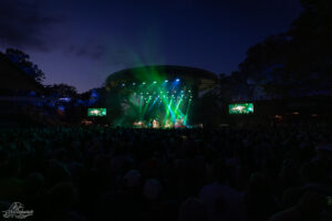 Outdoor concert at night with a large crowd facing a circular stage lit by green and blue lights, two side screens showing the performance