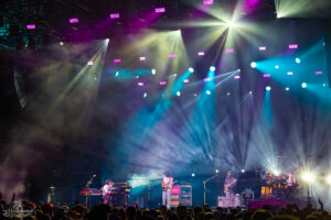 Live concert on a foggy stage with vibrant colored lights; keyboardist, guitarist, bassist, and drummer perform as the crowd watches.