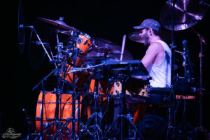 Drummer in a white sleeveless shirt and camo cap playing an orange drum kit on a dark stage with multiple cymbals and electronic pads nearby.