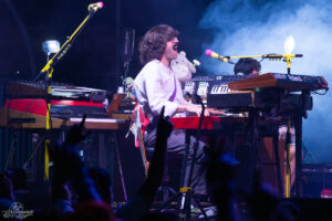Musician in white shirt singing into a mic while playing a red keyboard on a multi-synth stage; audience hands reach up in the foreground.