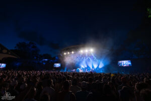 Outdoor night concert with a packed crowd facing a brightly lit circular stage and blue white spotlights cut through smoke.