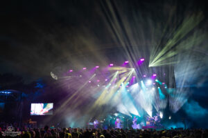 Outdoor night concert with a colorful light show and a crowd watching a band on stage; a large screen shows a guitarist on the left.
