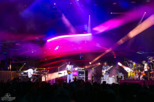 Live band performing on a brightly lit stage at night with pink, purple, and blue lights; guitarist center, keyboardist left, bassist right, drummer far right; crowd in foreground.