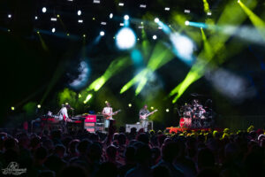 Band performing live on a dark stage under yellow and blue spotlights, crowded audience in the foreground posing the energy of a concert.