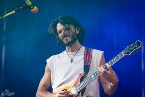 Male guitarist playing a wooden electric guitar on stage, wearing a white sleeveless shirt and necklace, with a blue smoky backdrop.