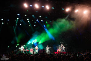 Band performing on a brightly lit stage with green and blue lights and a cheering crowd in the foreground.