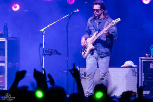 Bass guitarist on a blue-lit stage, wearing sunglasses and a denim jacket, as the crowd raises hands in front of him.