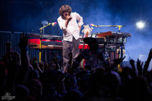 Musician in a white shirt performing on stage with multiple keyboards, waving to a cheering crowd amid blue stage lighting and smoke, a stuffed bunny sits on the gear nearby.